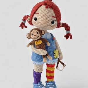 Könnte beinhalten: Handgefertigte Häkelpuppe mit roten Zöpfen, trägt ein blaues Kleid und blaue Schuhe. Die Puppe hält einen kleinen braunen Affen. Die Socken der Puppe sind lila und rot-gelb gestreift. Die Puppe hat einen gelben Blumenfleck auf dem Kleid.