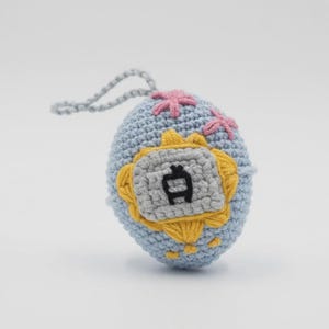 May include: A crocheted blue egg-shaped ornament with a gray and yellow emblem featuring a Japanese character. Pink flower-like accents are stitched on the top, and a gray loop is attached for hanging. The ornament is handmade.
