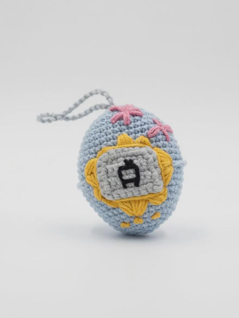 May include: A crocheted blue egg-shaped ornament with a gray and yellow emblem featuring a Japanese character. Pink flower-like accents are stitched on the top, and a gray loop is attached for hanging. The ornament is handmade.