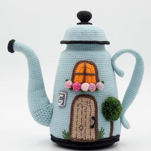 Puede incluir: Tetera de ganchillo azul claro con forma de casa, con puerta marrón, ventana naranja y un pequeño árbol verde. La tetera tiene un asa, un pico y una tapa negros, y está decorada con flores rosas y naranjas.