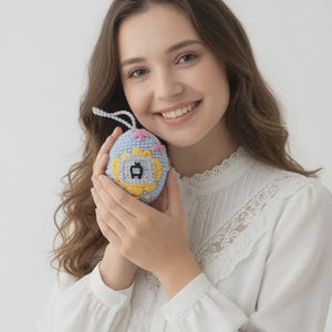 May include: A crocheted, light blue and yellow toy with a white string. The toy has a small screen with a yellow border and a black design. Pink and yellow flowers are crocheted on the top of the toy.