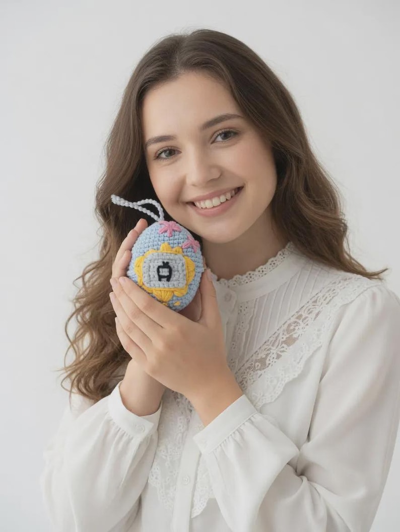 May include: A crocheted, light blue and yellow toy with a white string. The toy has a small screen with a yellow border and a black design. Pink and yellow flowers are crocheted on the top of the toy.