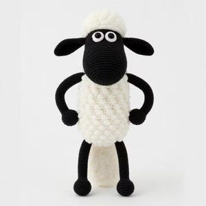 May include: A handmade crochet sheep toy with a white body, black legs, arms, and face, and a fluffy white head. The sheep has two small, round eyes and black ears. The toy is standing upright against a white background.
