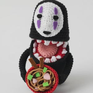 Puede incluir: Peluche de No-Face tejido a crochet, de El Viaje de Chihiro, con cara blanca, cuerpo negro y detalles morados. El peluche sostiene un cuenco de ramen tejido a crochet con fideos marrones, ingredientes verdes y rosas, y un cuenco rojo.