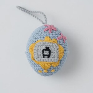May include: A crocheted egg-shaped ornament in light blue, featuring a gray and yellow emblem with a black symbol. Pink floral accents are stitched on top. A gray loop is attached for hanging.