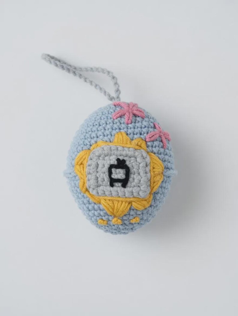 May include: A crocheted egg-shaped ornament in light blue, featuring a gray and yellow emblem with a black symbol. Pink floral accents are stitched on top. A gray loop is attached for hanging.