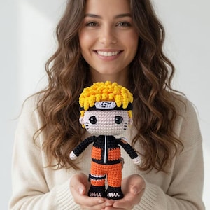 Pode incluir: Boneca de crochê feita à mão de um personagem com cabelo amarelo espetado, uma roupa preta e laranja e uma bandana. A boneca é segurada em frente a uma pessoa vestindo um suéter creme.