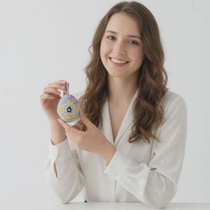 May include: A hand-stitched, egg-shaped ornament with a blue, yellow, and pink design. The ornament features a small house motif. The background is a plain white surface.