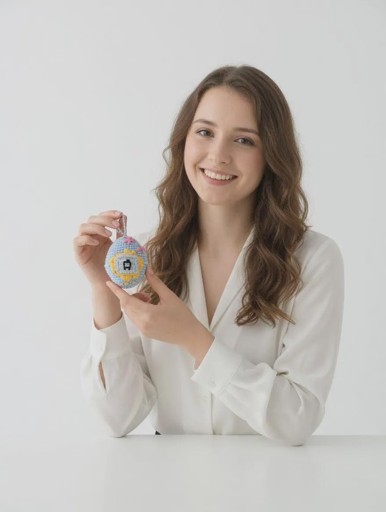 May include: A hand-stitched, egg-shaped ornament with a blue, yellow, and pink design. The ornament features a small house motif. The background is a plain white surface.