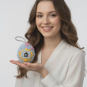 Puede incluir: Un juguete tejido en forma de huevo en azul claro, amarillo y rosa. El juguete presenta un pequeño gráfico gris y un lazo gris para colgar. El diseño recuerda a un juego portátil vintage. El juguete está en la mano de una persona.