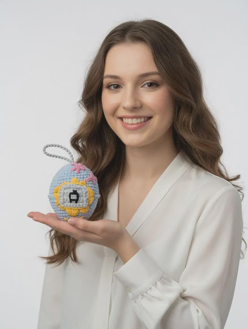 May include: A crocheted, egg-shaped toy in light blue, yellow, and pink. The toy features a small, gray graphic and a gray loop for hanging. The design is reminiscent of a vintage handheld game. The toy is held in a person's hand.