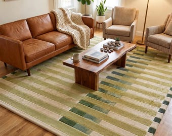 Peut inclure: Un grand tapis rectangulaire présentant un motif géométrique de rayures horizontales dans des tons variés de vert sauge, vert olive et crème. Le tapis possède un poil texturé et recouvre une partie importante du parquet du salon.