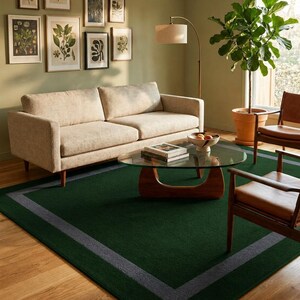 May include: A rectangular area rug featuring a deep forest green center field bordered by a thick, dark gray frame. The rug is positioned on a light-toned wooden floor, providing a solid foundation for the surrounding furniture and decor.