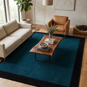May include: A rectangular area rug featuring a deep teal center panel framed by a wide, solid black border. The rug is positioned on a light-toned wooden floor, providing a bold color contrast to the surrounding furniture and decor items.