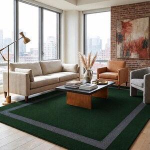 May include: A large rectangular area rug featuring a deep forest green center field framed by a wide, solid charcoal gray border. The rug has a low-pile, uniform texture and is positioned flat on a light-toned wooden floor surface.