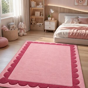 May include: A rectangular area rug featuring a light pink center panel framed by a darker pink scalloped border. The rug has a soft, textured surface and is positioned flat on a light-colored wooden floor, showcasing a clean, simple design.