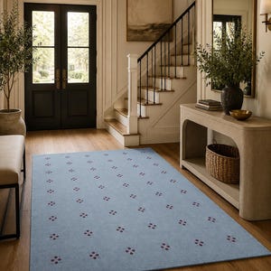 May include: A light blue rectangular rug with a repeating pattern of small, dark red floral-like designs. The rug is placed on a hardwood floor in a well-lit entryway, with a view of a staircase and a front door.