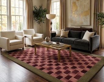 Peut inclure: Un tapis rectangulaire présentant un motif géométrique en damier répétitif dans des tons de bordeaux profond et de rose doux, encadré par une bordure vert olive unie. Le tapis est posé sur un parquet en bois sombre dans un salon.