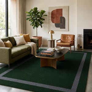 May include: A rectangular area rug featuring a deep forest green center field framed by a wide, solid charcoal gray border. The rug has a low-pile texture and is positioned on a smooth concrete floor, providing a dark foundation for the room.