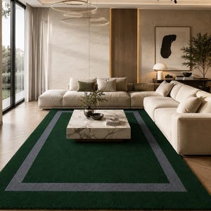 May include: A rectangular area rug featuring a deep forest green center field bordered by a wide, solid gray frame. The rug has a short, dense pile texture and is positioned on a light-toned wooden floor with a herringbone pattern in a room.