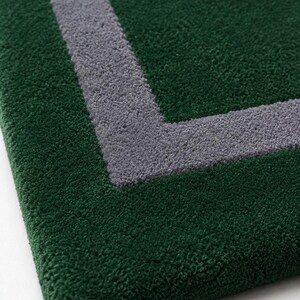 May include: A rectangular rug with a deep forest green pile and a contrasting thick gray border line forming a corner angle. The texture of the rug is dense and tufted, showing a clear distinction between the green and gray colored fibers.