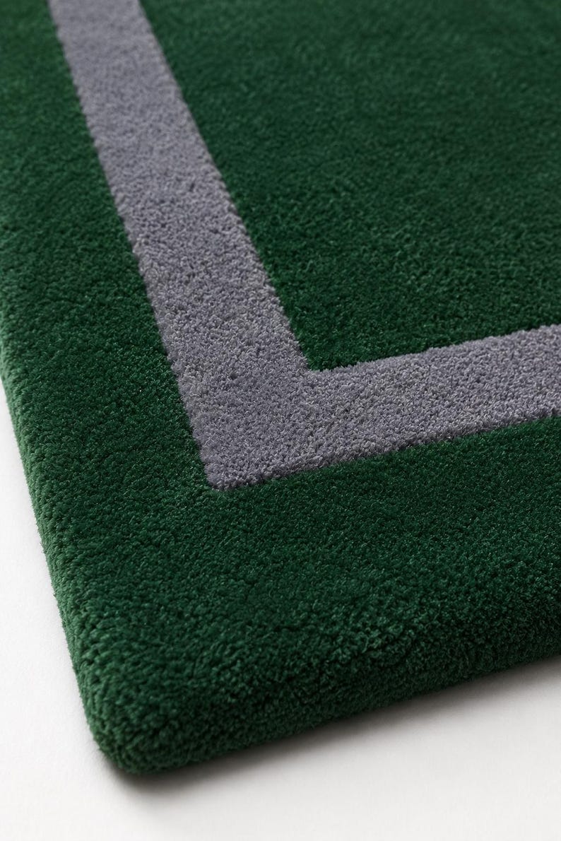 May include: A rectangular rug with a deep forest green pile and a contrasting thick gray border line forming a corner angle. The texture of the rug is dense and tufted, showing a clear distinction between the green and gray colored fibers.