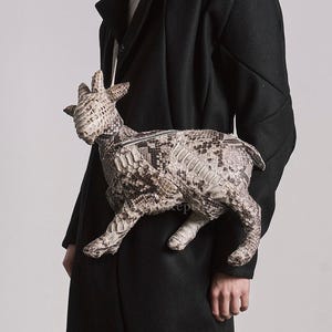 Puede incluir: Un bolso único con forma de cabra hecho de un material similar a la piel de serpiente. El bolso es principalmente beige y gris, con un patrón detallado. La persona lleva un abrigo largo negro y pantalones blancos.