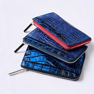 Puede incluir: Tres carteras rectangulares con textura de piel de cocodrilo azul y cremalleras de metal plateado. La cartera superior presenta un borde de tela roja brillante a lo largo de la cremallera. Cada cartera tiene un perfil delgado y reptiliano.