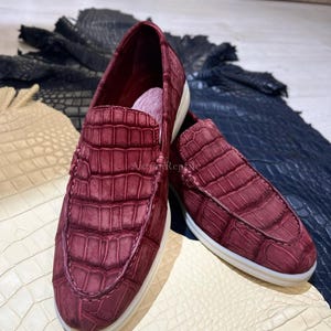 Puede incluir: Un par de zapatos sin cordones de cuero burdeos con textura de piel de cocodrilo. Los zapatos tienen suelas blancas y se exhiben sobre una superficie color crema. Los zapatos son de color rojo oscuro.