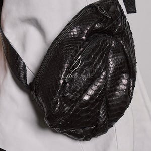 May include: Black leather crossbody bag with a textured, reptile-like pattern. The bag has a curved shape, a zippered main compartment, and a silver-toned zipper pull. The strap is black and made of the same material.