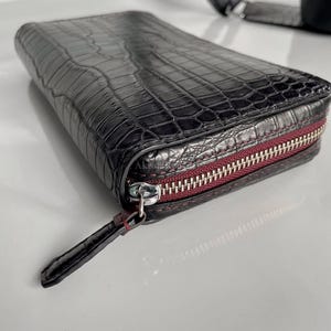以下が含まれることがあります： クロコダイル柄の型押しが施された長方形の黒い財布。シルバーカラーの金属製ファスナーと、同素材の黒いレザータブが付いています。ファスナーのテープ部分は深みのあるボルドーカラーで、コントラストを生み出しています。