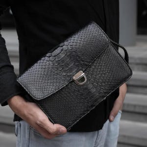 May include: Black leather clutch with a snakeskin texture. The clutch has a flap closure with a silver-colored clasp. A small handle is attached to the side. The clutch is held in front of a person.