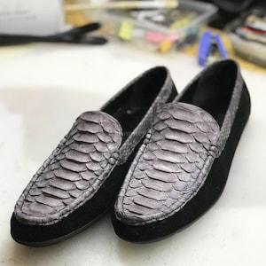 Pode incluir: Um par de mocassins sem atacadores com um padrão de pele de cobra cinzento texturizado no peito do pé e no calcanhar, contrastado por painéis laterais de camurça preta sólida e um design de biqueira arredondada. Silhueta de perfil baixo.