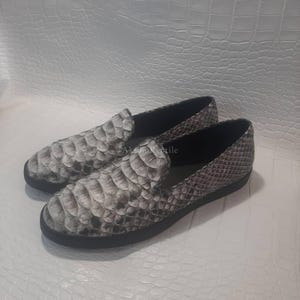 May include: A pair of slip-on shoes with a snakeskin pattern in shades of grey and white. The shoes have a rounded toe and a black sole. The texture of the snakeskin is visible, adding a unique detail to the footwear.