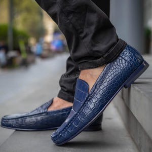 Puede incluir: Mocasines de cuero azul marino con textura de piel de cocodrilo. Estos zapatos sin cordones tienen un tacón bajo. La imagen muestra los zapatos en los pies de una persona, con la marca "Akrom Reptile" visible.