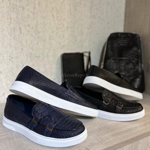 Peut inclure: Présentation de chaussures et d'accessoires de luxe. L'image présente deux paires de chaussures slip-on bleu marine avec des semelles blanches et des boucles, et une paire de chaussures noires. Un sac à dos et un portefeuille noirs sont également visibles.