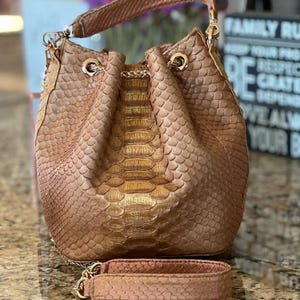 Op de afbeelding: Een bruin emmermodel tas met een bijpassende riem. De tas heeft een textuur, geschubd patroon met goudkleurige accenten. De tas heeft een trekkoordsluiting en goudkleurige hardware.