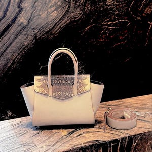 Può includere: Una borsa beige chiaro con manico superiore e tracolla staccabile. La patta presenta un motivo a pelle di serpente. La borsa è appoggiata su una superficie di marmo.