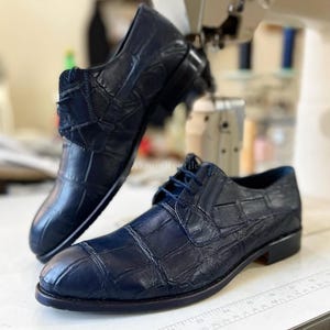 Puede incluir: Un par de zapatos de vestir de cuero azul marino con un patrón texturizado similar al de un cocodrilo. Los zapatos tienen cordones y un tacón bajo. La marca "AkromReptile" es visible en el fondo.