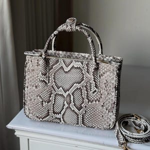 May include: A snakeskin-effect handbag with a top handle and a detachable shoulder strap. The bag has a zip closure and a neutral colour palette of white, grey, and black. The bag is on a white surface.