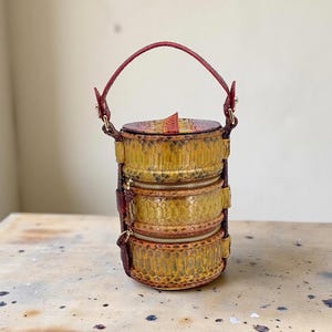 May include: A cylindrical handbag with three zippered compartments, crafted from a snakeskin-like material in shades of yellow, brown, and red. It features a red handle and accents, and a top flap.