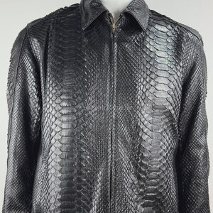 Könnte beinhalten: Eine schwarze Jacke mit einem strukturierten Schlangenledermuster auf dem gesamten Kleidungsstück. Die Jacke verfügt über einen klassischen Kragen, lange Ärmel und einen zentralen Reißverschluss auf der Vorderseite.