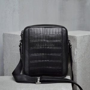 May include: Black crossbody bag with a textured crocodile pattern. The bag has a rectangular shape with a long adjustable strap. The brand name "AkromReptile" is visible on the front of the bag.