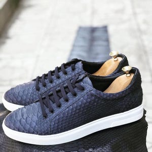 May include: A pair of low-top sneakers featuring a dark navy blue textured scale pattern on the upper, black laces, and a solid white rubber sole. Each shoe contains a light-colored wooden shoe tree insert with a gold-toned metal knob at the heel.