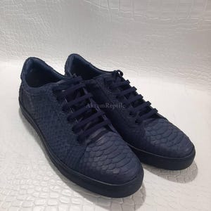 Puede incluir: Un par de zapatillas bajas de color azul marino con un patrón de escamas texturizado en la parte superior, cordones azul marino a juego y una suela de goma azul marino. El texto AkromReptile es visible en el lateral de la zapatilla.