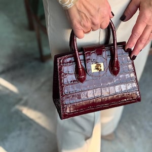 Puede incluir: Un bolso de mano de color burdeos con textura de piel de cocodrilo. El bolso tiene forma rectangular, con un asa superior y un cierre dorado. El bolso lo sostiene una persona que lleva una parte superior y pantalones beiges.