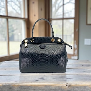 May include: Black leather handbag with a snakeskin pattern. The bag features a top handle, a gold zip, and gold-coloured hardware. The bag is on a wooden table in front of a window.