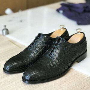 May include: A pair of black leather dress shoes with a textured, reptile-like pattern. The shoes have blue laces and are displayed on a light-coloured surface. The shoes have wooden shoe trees inside.