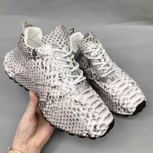 May include: A pair of low-top sneakers featuring a textured snakeskin pattern in shades of white, gray, and black. The shoes have white laces, a white interior lining, and a black rubber outsole. The exterior material displays a repeating scale design.