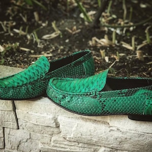 May include: A pair of green leather loafers with a snakeskin pattern. The shoes are a vibrant green color and have a classic slip-on design. The loafers are displayed on a stone wall with a blurred background of greenery.